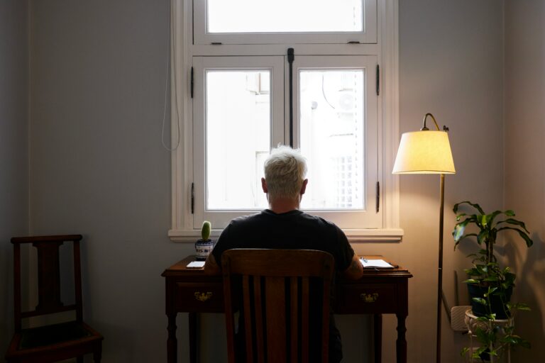 Older adult with white hair sitting alone at a wooden desk, facing a window in a dimly lit room